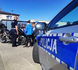 obraz przedstawia policjanta z dziećmi na placu komendy, w tle radiowóz i motocykl