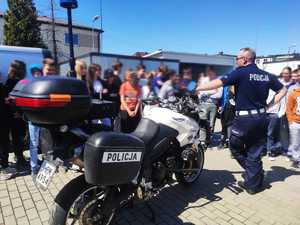 obraz przedstawia policjanta z młodzieżą na placu komendy, w tle radiowóz i motocykl