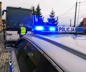 obraz przedstawia policjanta sprawdzającego autokar, w tle radiowóz