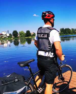 obraz przedstawia policjanta na rowerze  na tle zbiornika wodnego
