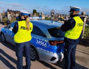 obraz przedstawia policjantów patrzących na cmentarz na tle radiowozu